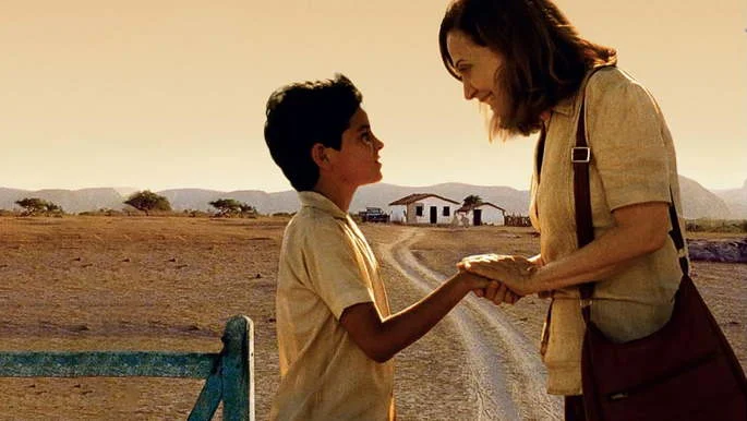 Um menino com cerca de 10 anos e uma mulher de cerca de 50 anos com as mãos dados, se olhando em meio a uma paisagem do sertão nordestino.