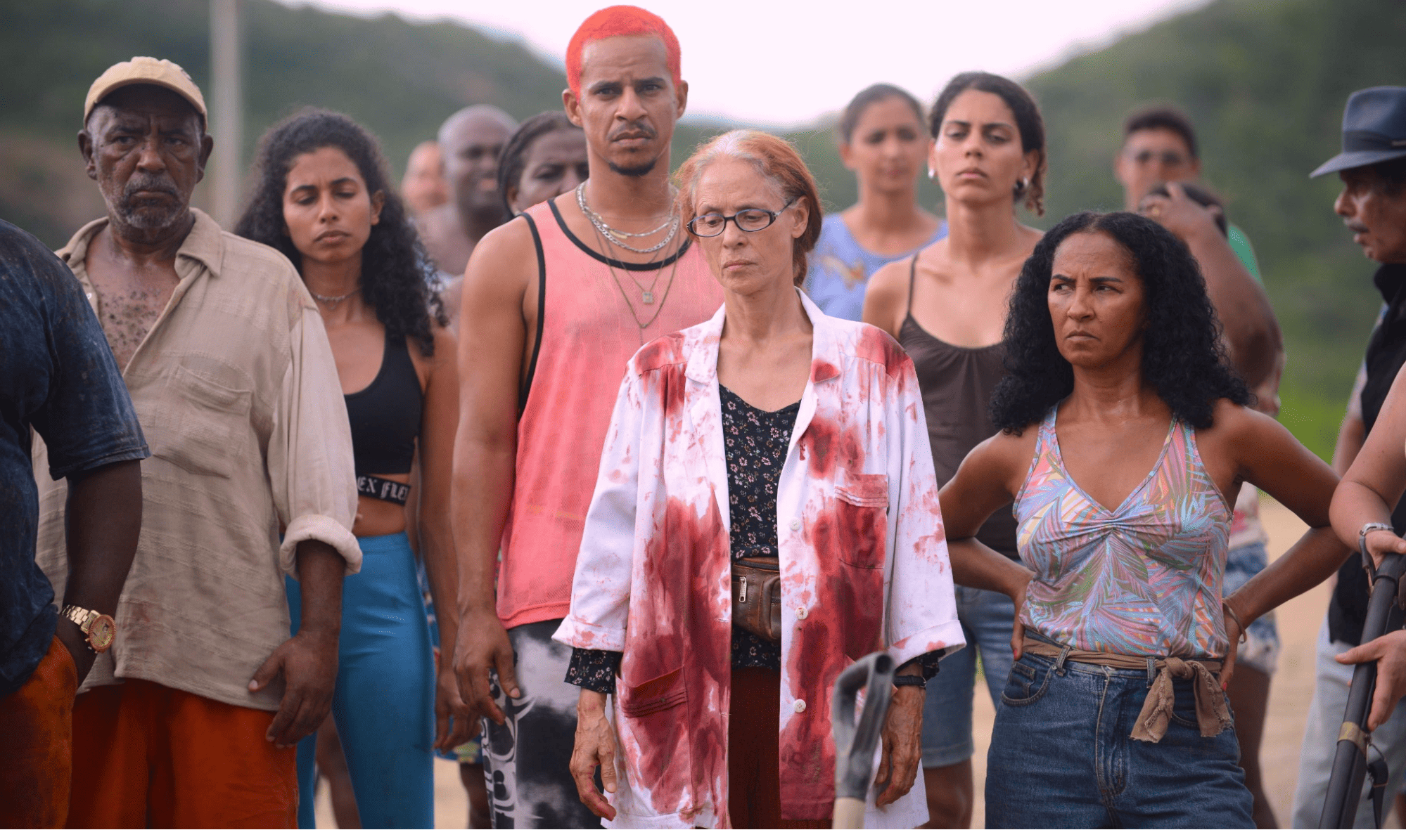 Um grupo de pessoas reunidas olhando para algo. A mulher em destaque está com a roupa suja de um líquido vermelho.